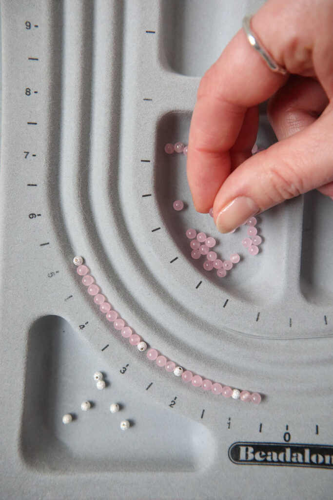 Minuscules perles de 4 mm en argent et rose sur un tableau de perles grises, avec la main d'une femme blanche créant un motif de bijoux