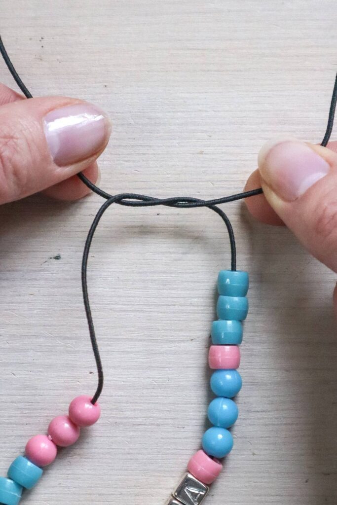 Comment faire un bracelet en perles - mains attachant l'élastique en prenant l'extrémité gauche de l'élastique par-dessus, derrière et devant la droite