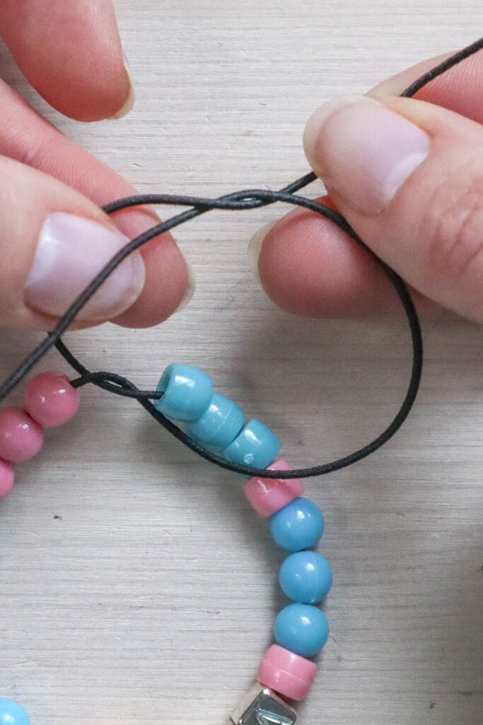 Comment faire un bracelet en perles - mains attachant l'élastique en prenant l'extrémité droite de l'élastique par-dessus, derrière et devant la gauche