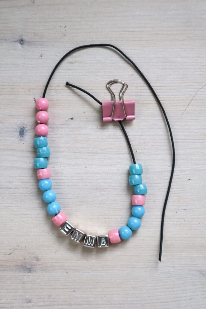 Comment fabriquer un bracelet de perles : des perles élastiques bleues et roses enfilées sur une courte longueur d'élastique en tissu noir, avec une pince-reliure fixant une extrémité. 